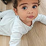 baby, child, curly_hair, cute, expression, face, floor, hands, indoor, large_eyes, laying_down, long_sleeves, looking_up, person, portrait, skin, toddler, white_clothing, wooden_floor, young_child