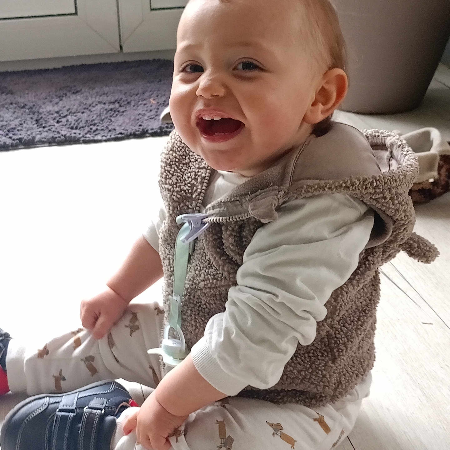 Mylan participe au concours pour gagner de l'argent avec cette photo : baby, child, clothing, cute, dog_pattern, floor, happy, home, indoor, light, pacifier, pants, plant_pot, rug, shirt, shoes, sitting, smiling, toddler, vest