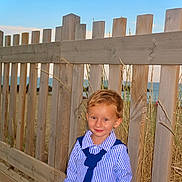 Conor participe au concours pour gagner de l'argent avec cette photo : child, boy, smile, striped_shirt, tie, wooden_fence, beach, ocean, sky, grass, sand, outdoor, portrait, casual_clothing, sunlight, young_child, nature, happy, standing, blond_hair