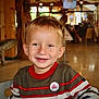 child, boy, smiling, indoor, sweater, striped_clothing, blond_hair, happy, portrait, casual_wear, face, seated, young, cute, warm_lighting, wooden_interior, restaurant, background_blur, person, kid