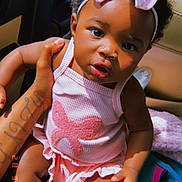 Nyari is registered to the contest to win money with this photo: baby, child, pink_bow, pink_outfit, car_seat, tattoo, hand, curly_hair, infant, cute, portrait, person, indoors, seat, clothing, headband, skin, expression, holding, young_child