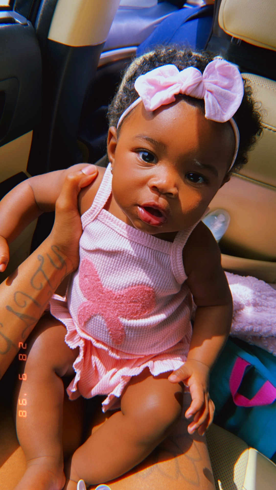 Nyari is registered to the contest to win money with this photo: baby, child, pink_bow, pink_outfit, car_seat, tattoo, hand, curly_hair, infant, cute, portrait, person, indoors, seat, clothing, headband, skin, expression, holding, young_child