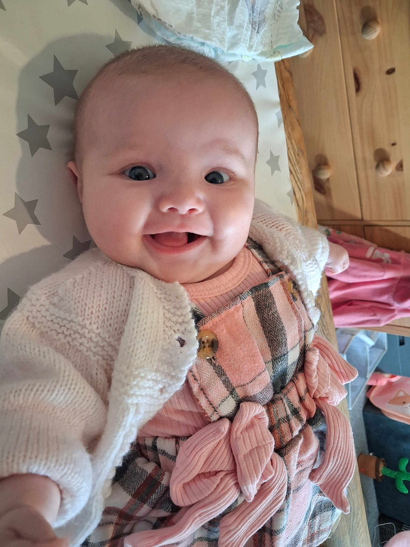 Emma-Leigh joined the competition — help win amazing prizes! baby, smiling, child, infant, knitted_cardigan, pink_dress, plaid_pattern, bows, happy, face, lying_down, stars_pattern, wooden_furniture, indoor, cute, person, apparel, toy, room, candid