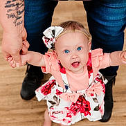 Harper is registered to the contest to win money with this photo: baby, child, blue_eyes, dress, floral, headband, holding_hands, infant, joyful, person, standing, wooden_floor, adult_hands, tattoos, happy, cute, family, support, indoors, footwear