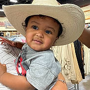 Kyzen is registered to the contest to win money with this photo: toddler, child, cowboy_hat, indoor, clothing_store, person, holding, curly_hair, short_sleeve_shirt, gray_shirt, bracelet, adult_hand, shopping, hanging_clothes, rack, casual, portrait, cute, bright_lighting, curious