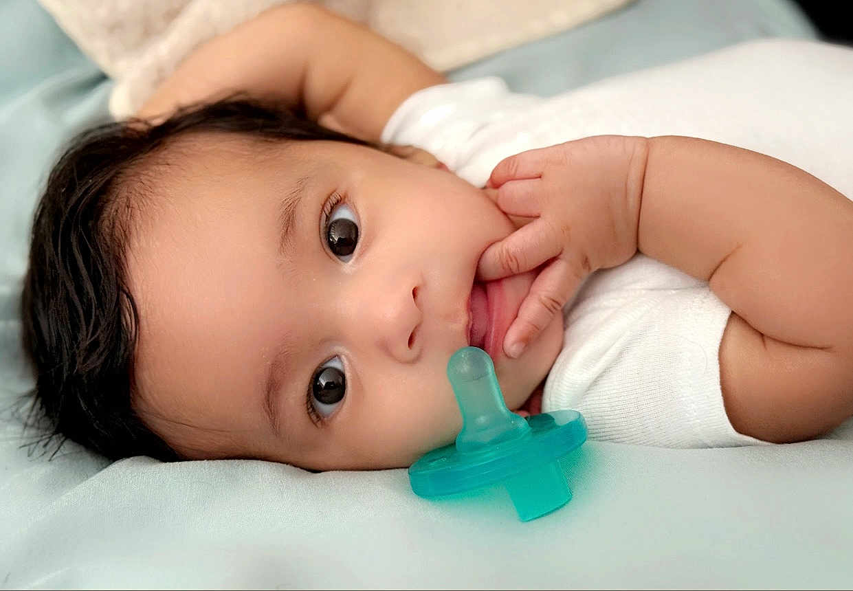 Brynn joined the competition — help win amazing prizes! baby, infant, pacifier, hand, face, eyes, skin, lying_down, white_clothing, soft_surface, curly_hair, closeup, cute, child, newborn, portrait, fingers, sleepwear, resting, indoors