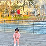 child, toddler, pink_clothing, walking, boardwalk, water_fountain, outdoor, daylight, trees, river, railing, building, person, footwear, casual_wear, nature, young_child, scenic, sunlight, park