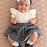 baby, infant, child, portrait, smiling, lying_down, headband, flower, ruffled_blouse, skirt, houndstooth, dress_shoes, bows, socks, quilted_blanket, pink_background, indoor, fashion, cute, photography