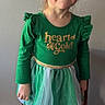child, girl, green_dress, bow, boots, standing, chair, smiling, indoor, portrait, happy, clothing, fashion, footwear, long_sleeves, tights, wall, floor, person, cute