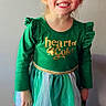 child, girl, smiling, green_dress, bow, boots, standing, chair, happy, portrait, indoor, long_sleeves, tulle_skirt, gold_text, light_hair, casual, cute, fashion, wall, floor