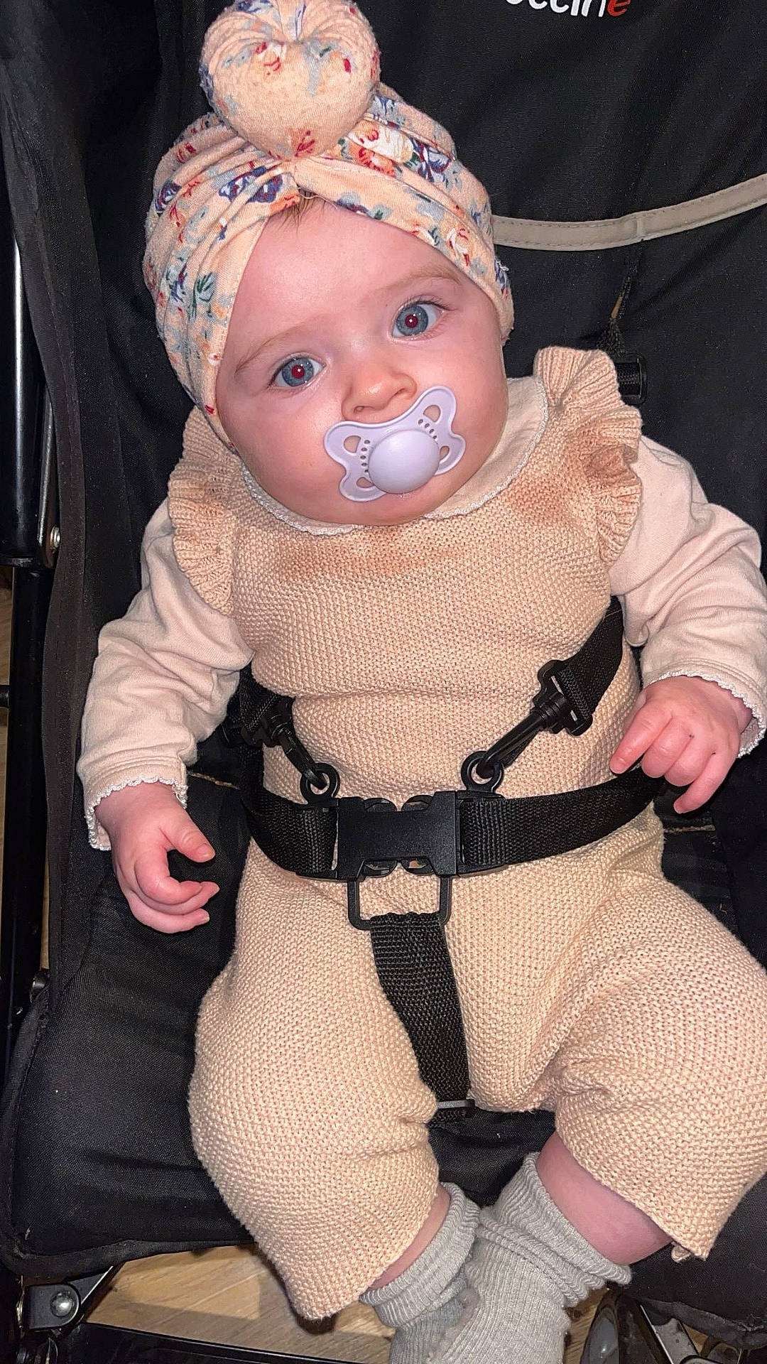 baby, buckle, child, closeup, eyes, face, hands, headwrap, indoor, infant, knitted_outfit, onesie, pacifier, portrait, red_eye, safety_belt, sitting, socks, stroller, wooden_floor