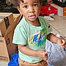 Kordell is registered to the contest to win money with this photo: brown_pants, cardboard_boxes, casual_clothing, child, curly_hair, expression, floor_tiles, footwear, green_shirt, home_interior, indoor, looking_at_camera, person, shoe_rack, shoes, standing, storage, striped_socks, toddler, young_child