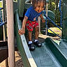 black_shoes, blue_shirt, braided_hair, child, daytime, handrail, happy, outdoor, park, person, play_equipment, playground, recreation, red_shorts, slide, smiling, socks, summer, sunlight, trees