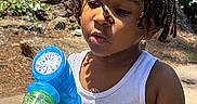 Kordell joined the competition — help win amazing prizes! child, boy, braided_hair, bubble_gun, bubbles, outdoor, sunlight, white_tank_top, nature, greenery, rocks, tree_trunk, playing, summer, daylight, portrait, curious, hands, expression, young