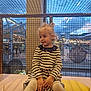 toddler, child, blonde_hair, striped_dress, sitting, cushion, netting, indoor, evening, lights, urban, window, play_area, cute, portrait, small_hands, white_tights, casual_clothing, young_girl, smiling