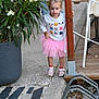 child, toddler, girl, pink_tutu, sweatshirt, animal_print, sidewalk, plant, flowers, wooden_post, outdoor, curious, shoes, rope, potted_plant, concrete, casual, daylight, cute, standing