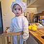 child, toddler, chef_hat, apron, flour, kitchen, lemons, wooden_chair, countertop, cabinet, sink, person, cooking, indoors, cute, surprised, blonde_hair, white_clothing, floor_tiles, home
