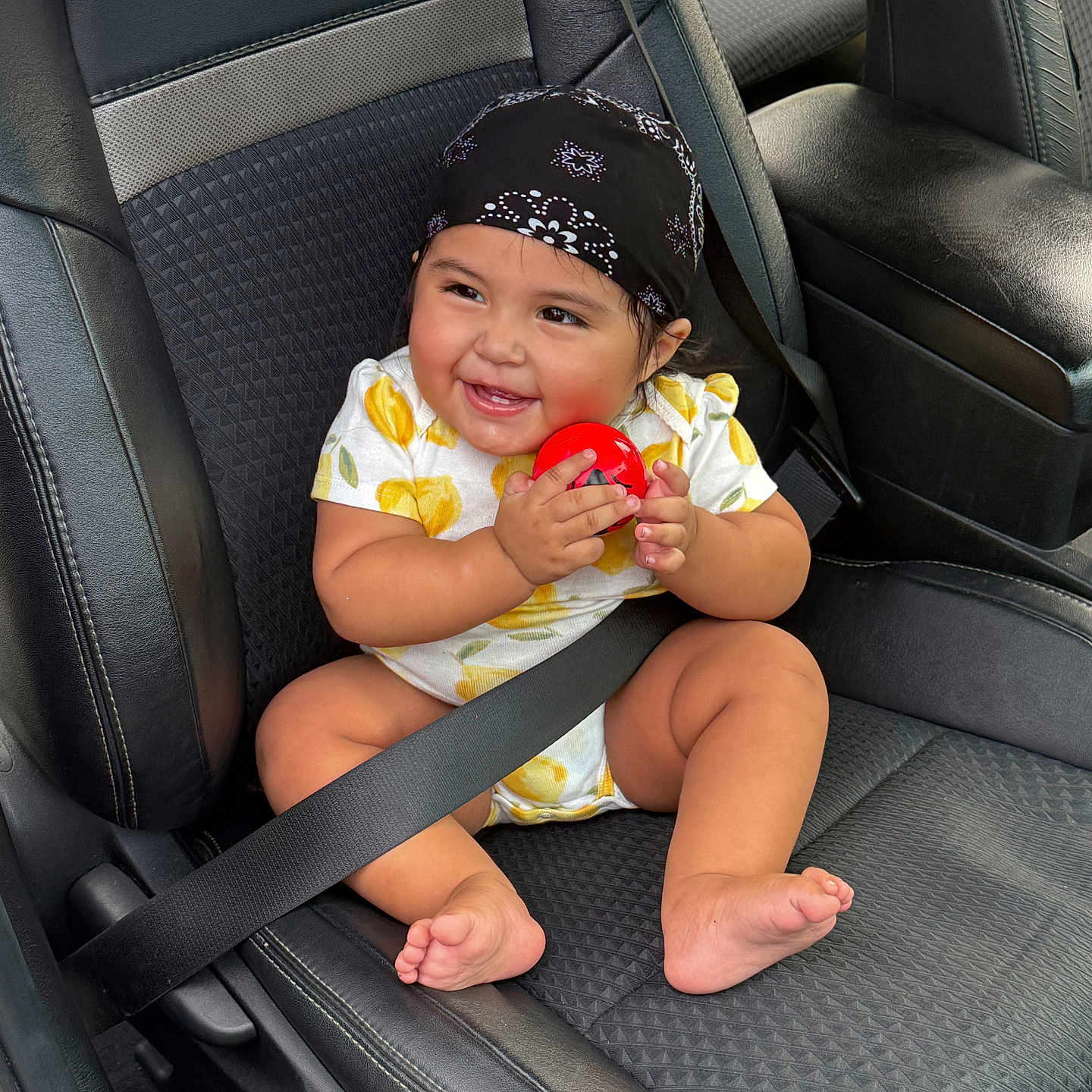 Megan is registered to the contest to win money with this photo: baby, bandana, black_leather, car_seat, child, clothing, cute, foot, hand, happy, headwear, indoors, infant, lemon_pattern, person, red_ball, seatbelt, sitting, smiling, toy