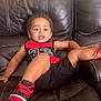 toddler, child, leather_couch, red_shoes, black_shorts, red_and_black_shirt, sitting, smiling, indoor, casual, playful, curly_hair, footwear, relaxed, young_child, comfortable, furniture, portrait, happy, expression