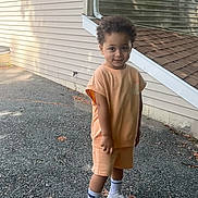 Kingston is registered to the contest to win money with this photo: child, boy, curly_hair, smiling, outdoor, house, wall, sidewalk, sneakers, nike, shorts, tshirt, standing, daylight, shadow, concrete, casual_clothing, cute, young, portrait