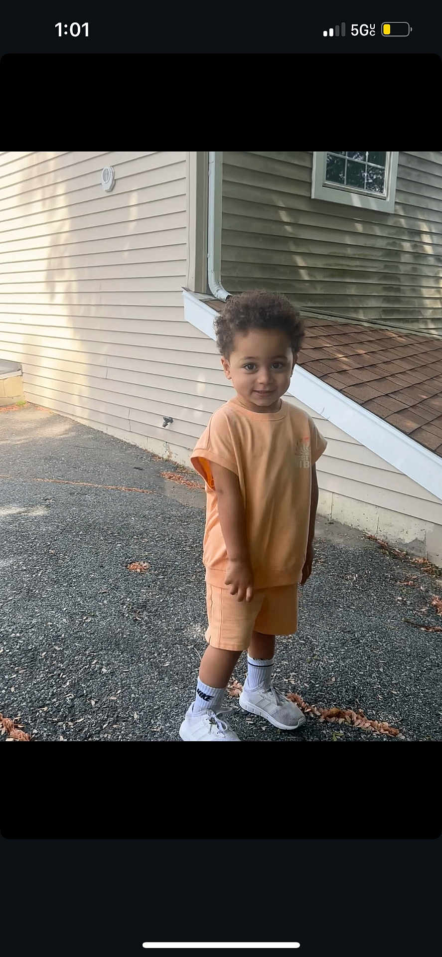 Kingston is registered to the contest to win money with this photo: child, boy, curly_hair, smiling, outdoor, house, wall, sidewalk, sneakers, nike, shorts, tshirt, standing, daylight, shadow, concrete, casual_clothing, cute, young, portrait