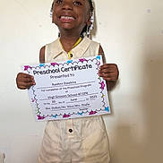 Raeann joined the competition — help win amazing prizes! achievement, celebration, certificate, child, clothing, education, face, hair_beads, hands, happy, indoor, person, portrait, preschooler, proud, shorts, smiling, standing, wall, young