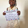 achievement, celebration, certificate, child, clothing, education, face, hair_beads, hands, happy, indoor, person, portrait, preschooler, proud, shorts, smiling, standing, wall, young