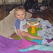 Blake joined the competition — help win amazing prizes! toddler, child, smiling, indoor, toy, blanket, playmat, floor, pillow, backpack, chair, person, clothing, wood_floor, window, adult_arm, casual, happy, candid, home