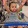 toddler, child, shopping_cart, pacifier, grocery_store, aisle, person, clothing, dress, hair_bow, hand_raised, shopping, retail, supermarket, food, snack, background, store_shelf, shopping_basket, happy