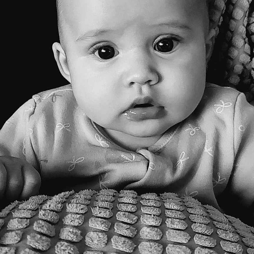 Margot-Nevaeh is registered to the contest to win money with this photo: baby, black_and_white, blanket, close_up, clothing, curious, cute, expression, eyes, face, head, indoors, infant, monochrome, person, portrait, sitting, soft_light, texture, young_child