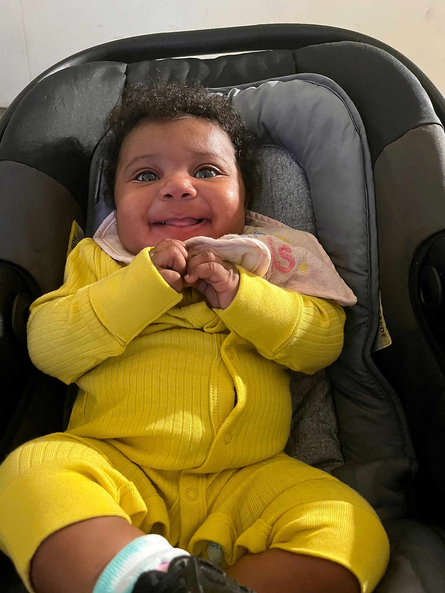 Tyla is registered to the contest to win money with this photo: baby, child, smiling, yellow_clothing, car_seat, happy, infant, cute, hands, face, curly_hair, indoor, person, seat, young_child, cozy, portrait, sitting, apparel, closeup