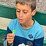 child, boy, bench, whipped_cream, dessert, food, green_spoon, outdoor, casual_clothing, blue_sweater, face, expression, eating, close_up, daylight, park, young, person, nose, hand