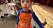 Conrad is registered to the contest to win money with this photo: child, boy, orange_apron, hardware_store, wood_planks, tool, backpack, water_bottle, people, indoor, curious, wide_eyes, floor, clothing, sneakers, table, paint_stains, adult, shopping, busy