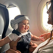 Adrian is registered to the contest to win money with this photo: baby, headphones, smile, airplane, window, adult, holding, overalls, white_shirt, happy, child, seat, travel, person, glasses, light, inside, portrait, interaction, cute