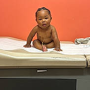 Knowledge is registered to the contest to win money with this photo: baby, child, medical_table, orange_wall, gold_chain, sitting, curious, indoor, person, skin, hair, expression, looking, hands, feet, clinic, healthcare, furniture, wall, portrait