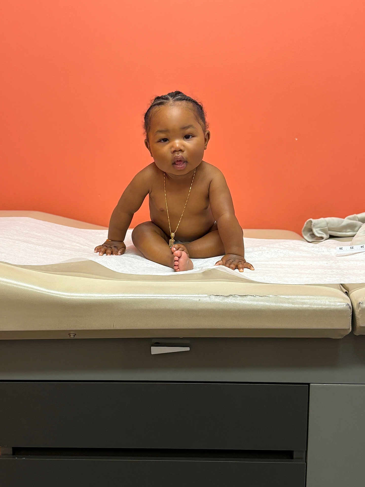 Knowledge is registered to the contest to win money with this photo: baby, child, medical_table, orange_wall, gold_chain, sitting, curious, indoor, person, skin, hair, expression, looking, hands, feet, clinic, healthcare, furniture, wall, portrait