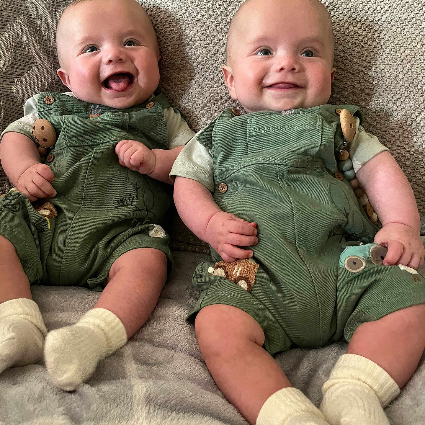 Tate And Trent is registered to the contest to win money with this photo: baby, blanket, child, close_up, couch, cushion, cute, face, green_overalls, happy, indoor, infant, joy, person, playful, portrait, sitting, smile, two_babies, white_socks