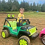 child, toddler, toy_car, jeep, outdoor, muddy, grass, dirt, yellow_shirt, green, play, vehicle, nature, trees, sky, cloudy, wheels, park, serious, boy