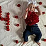 baby, infant, child, red_roses, white_blanket, bow, headband, floral_top, dark_pants, barefoot, smiling, happy, portrait, laying_down, cute, celebration, flower_arrangement, soft_texture, indoor, closeup