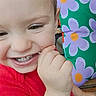 brown_hair, child, close_up, colorful, cushion, daylight, face, finger_biting, flower_pattern, happy, outdoor, person, photography, playful, portrait, red_shirt, seated, smiling, teeth, toddler