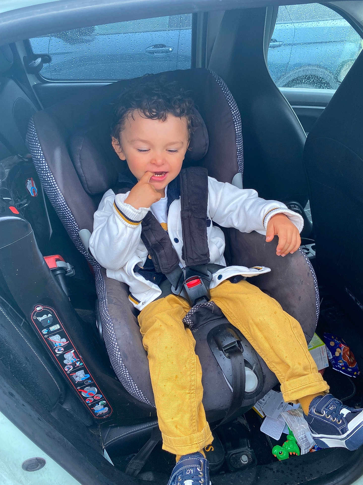 Ismaël participe au concours pour gagner de l'argent avec cette photo : child, toddler, car_seat, car_interior, curly_hair, white_jacket, yellow_pants, blue_shoes, rain_drops, window, seat_belt, vehicle, playful, indoors, seat, clothing, person, smiling, casual, messy_floor