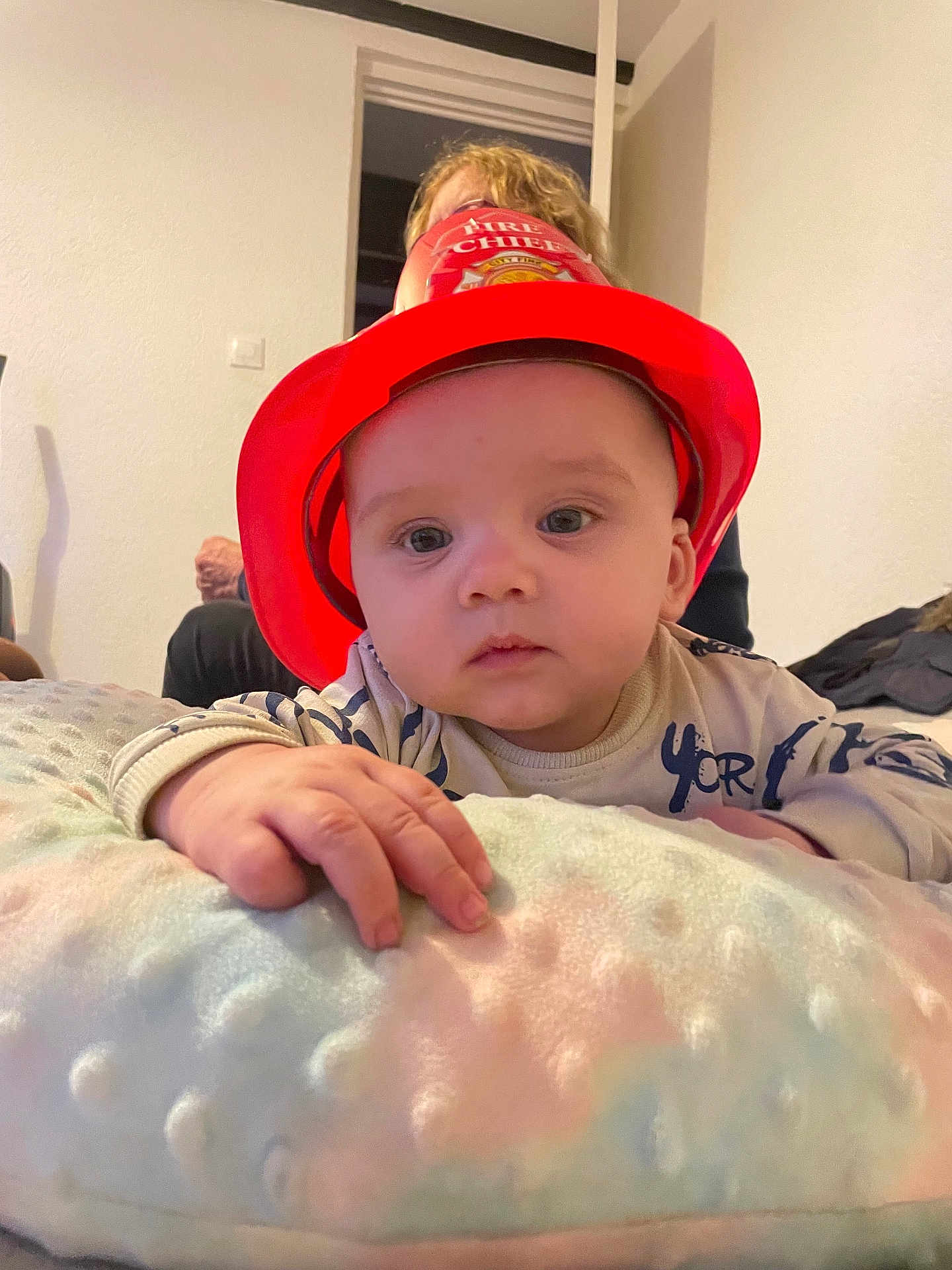 Ethan a rejoint le concours — aidez-le/la à gagner de superbes lots ! baby, child, fire_chief_hat, red_hat, blanket, indoor, person, hand, curious, soft_texture, blonde_hair, adult, wall, light, reaching, cute, infant, young_child, portrait, cozy