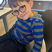 Amir joined the competition — help win amazing prizes! child, boy, portrait, smiling, glasses, eyewear, striped_shirt, blue_clothing, gray_clothing, seated, indoor, optical_shop, eyeglasses_store, chair, furniture, counter, store, short_hair, face, photography
