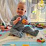 baby, child, colorful, cute, daylight, face, floor, footwear, hands, happy, indoor, infant, pajamas, plastic_toys, playmat, playpen, room, sitting, smiling, toys