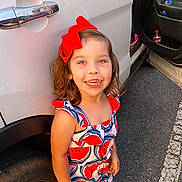 Gracie is registered to the contest to win money with this photo: car, child, cute, daylight, fashion, girl, happy, outdoor, parking_lot, person, red_bow, road, shorts, sleeveless, smiling, summer_clothing, tire, watermelon_print, wheel, white_car