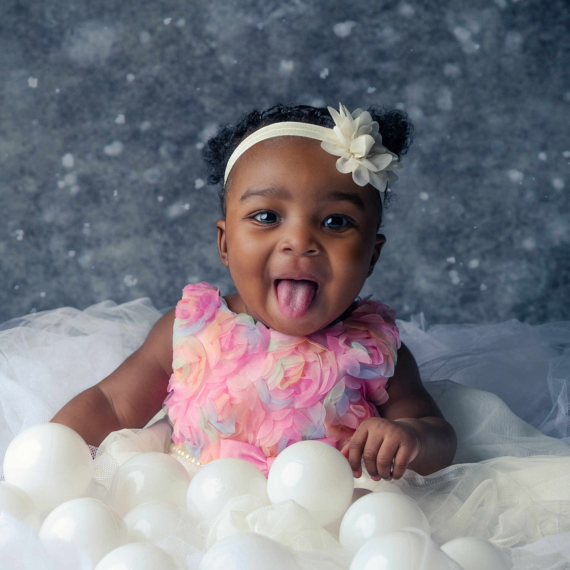 Jasmine is registered to the contest to win money with this photo: baby, child, girl, smiling, tongue_out, afro_puffs, headband, flower, pink_dress, white_balls, tulle, fabric, portrait, playful, cute, studio, background, happy, sitting, person