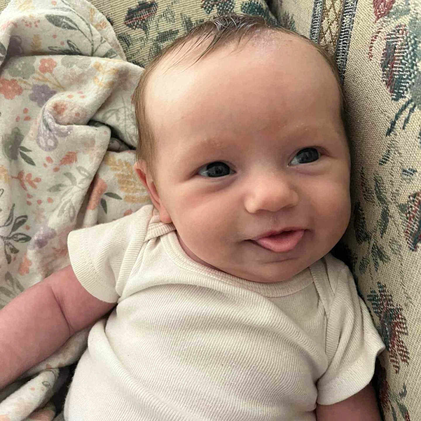 Emileigh is registered to the contest to win money with this photo: baby, blanket, cheeks, clothing, comfort, couch, cute, eyes, fabric, floral_pattern, head, indoors, infant, person, portrait, relaxing, skin, sleeve, smiling, tongue_out