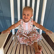Paisley is registered to the contest to win money with this photo: child, toddler, girl, smiling, happy, portrait, sitting, rocking_chair, barefoot, dress, floral_dress, outdoor, porch, blue_wall, wooden_chair, white_chair, summer, lifestyle, single_subject, photography