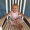 child, toddler, girl, smiling, happy, portrait, sitting, rocking_chair, barefoot, dress, floral_dress, outdoor, porch, blue_wall, wooden_chair, white_chair, summer, lifestyle, single_subject, photography