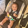 baby, car_seat, smiling, happy, infant, pacifier, snack_pouch, legs, feet, clothing, indoor, floor, bag, tiled_floor, child, person, seatbelt, cute, expression, holding
